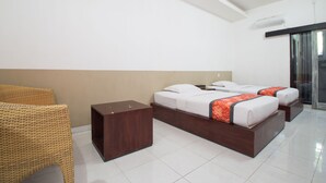 A modern hotel room with two beds, a wooden nightstand, a wicker chair, and a glass door leading to a balcony. - Adikara Renon (Denpasar)