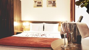 Una habitación de hotel con una cama, dos cuadros en la pared, una mesita de noche de madera con dos vasos, y un florero con flores.