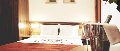 A hotel room with a bed, two framed pictures on the wall, a wooden nightstand with two glasses, and a vase with flowers.