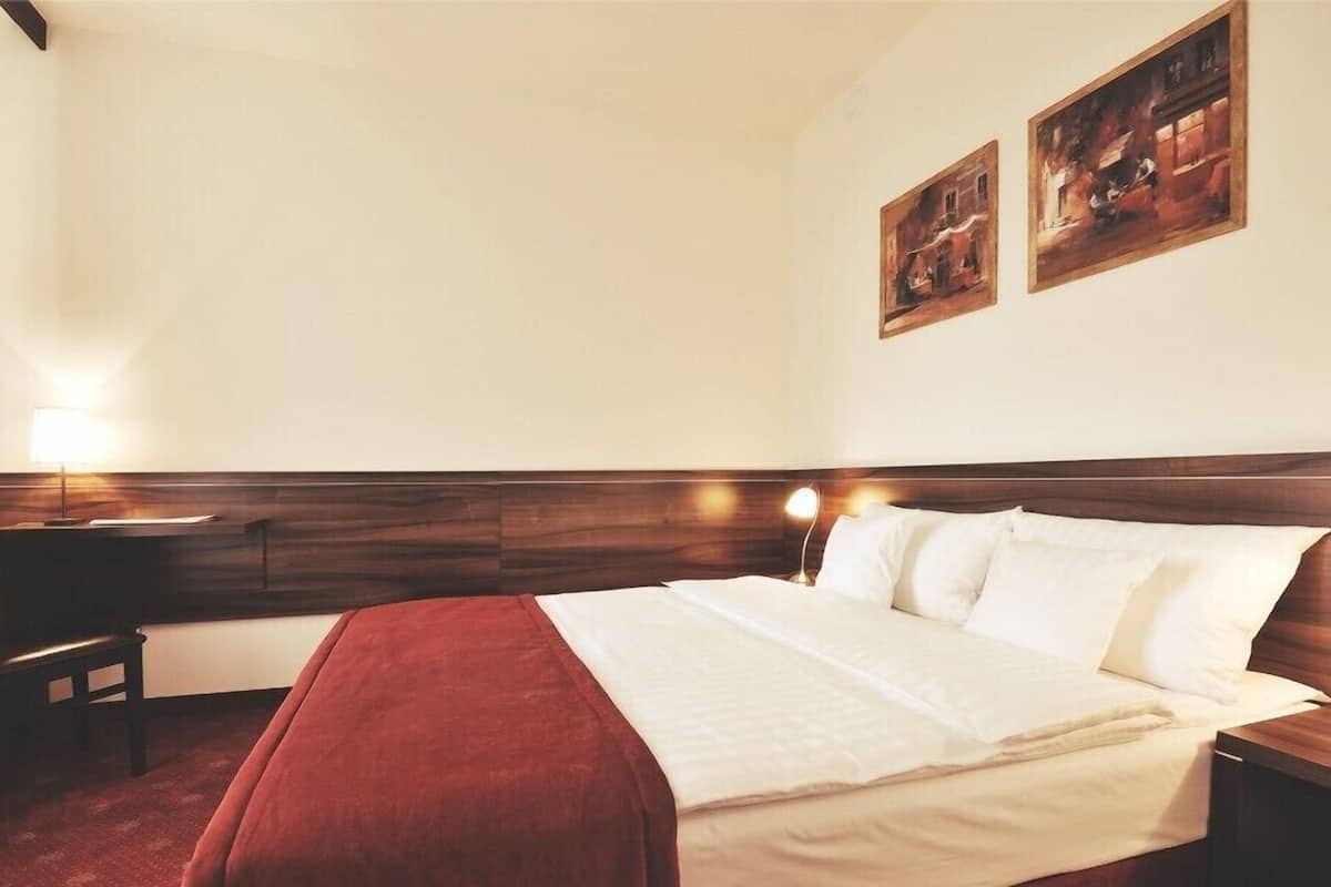 A hotel room with a wooden headboard, a bed with white linens, a red throw blanket, two bedside tables, and two wall-mounted paintings.