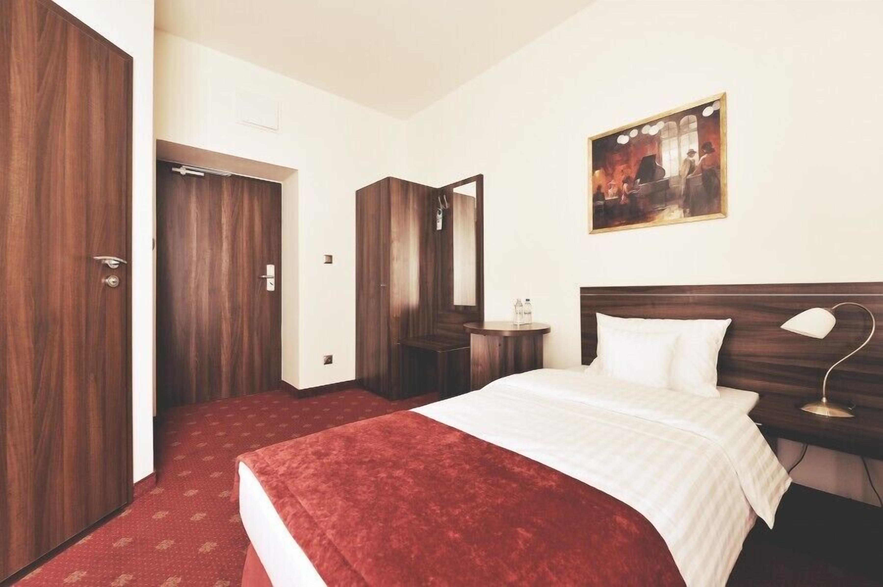 A hotel room with a large bed, a wooden wardrobe, a bedside table with a lamp, and a painting on the wall.