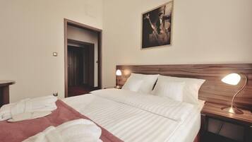 Una camera d'albergo con un letto grande, due lampade ai comodini, una testiera in legno, un quadro appeso al muro e una porta che dà su un'altra stanza.