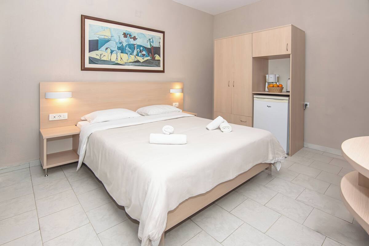 A neatly arranged hotel room with a large bed, bedside tables with lamps, a small kitchenette, and a painting on the wall.