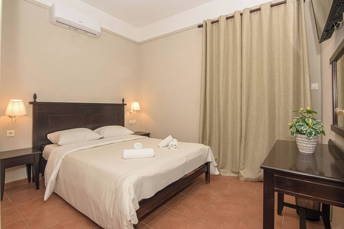 A hotel room with a bed, two bedside lamps, a desk, a potted plant, and a window with curtains.