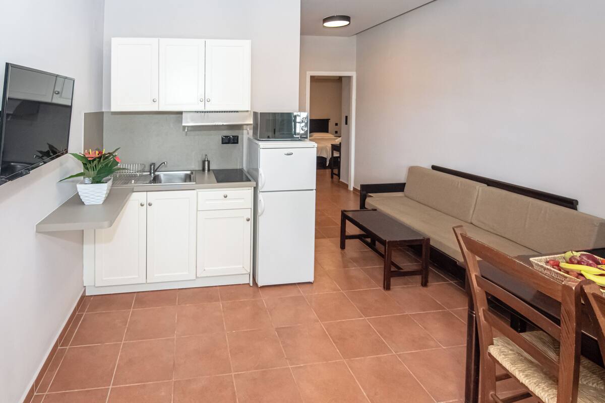 A compact kitchen with white cabinets, a microwave, and a refrigerator. There is a dining area with a wooden table and chairs, and a sofa.