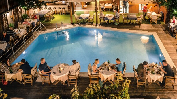 Una zona de menjador a la piscina amb taules i cadires disposades al voltant, envoltada d'un terra de fusta i vegetació.