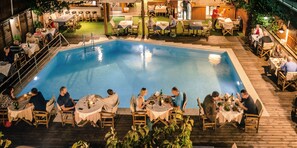 A poolside dining area with tables and chairs arranged around the pool, surrounded by wooden decking and greenery.