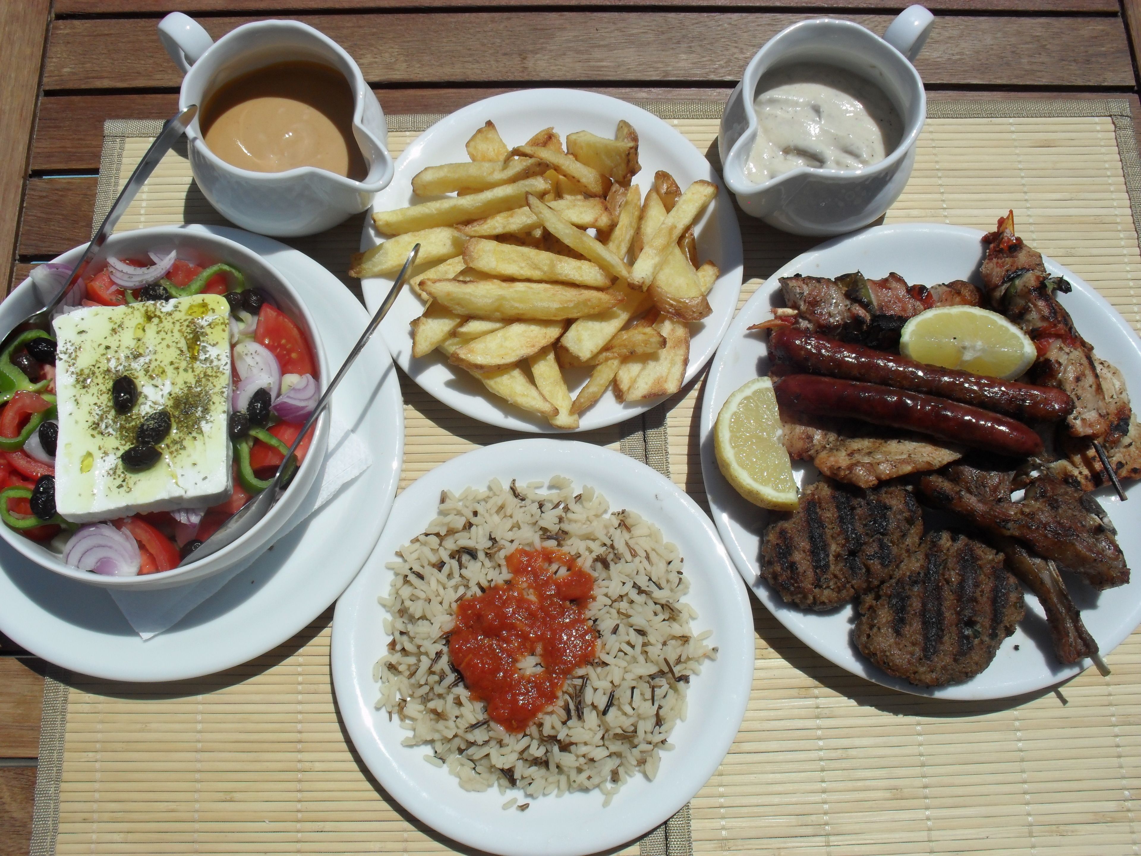 Une table garnie de différents plats, notamment une salade, des frites, du riz à la sauce tomate, de la viande grillée et des saucisses.
