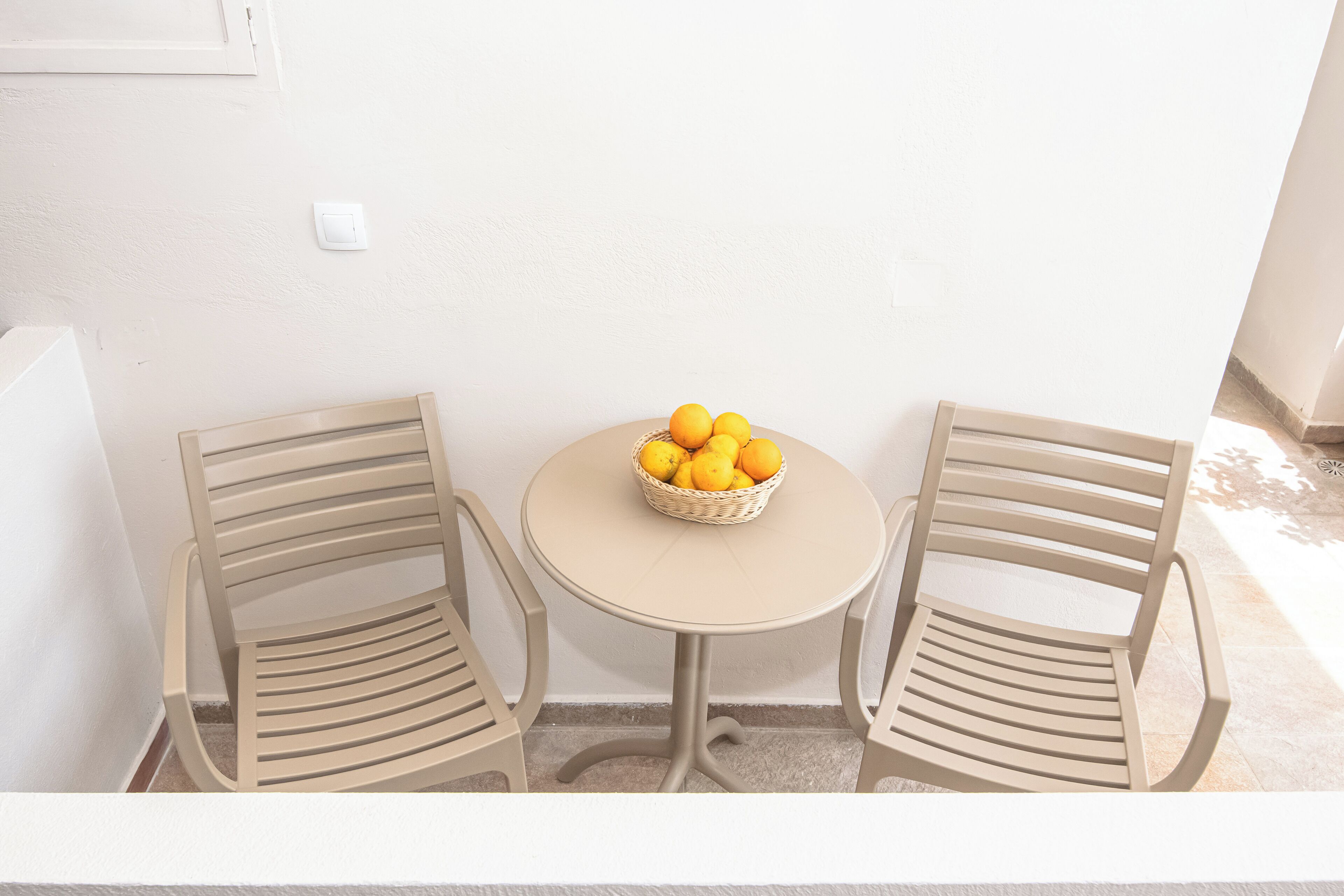 A small outdoor seating area with two beige chairs and a round table, featuring a basket of oranges.