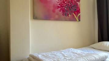 Una cama bien hecha con edredón blanco y almohadas, un cuadro colgado en la pared con flores rosas y una cortina oscura a la derecha.