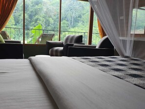 A bedroom with a large bed, two armchairs, and a view of a lush forest through large windows. - Ayubo Ella - Bed & Breakfast (Ella)