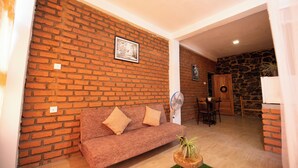 A living room with a brick wall, a sofa with cushions, a small table with a plant, a fan, and a dining area with chairs. - Ayubo Ella - Bed & Breakfast (Ella)