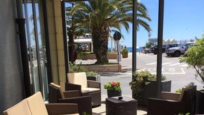 An outdoor seating area with wicker furniture, potted plants, and a view of the street and palm trees. - Hotel Internazionale (Finale Ligure)
