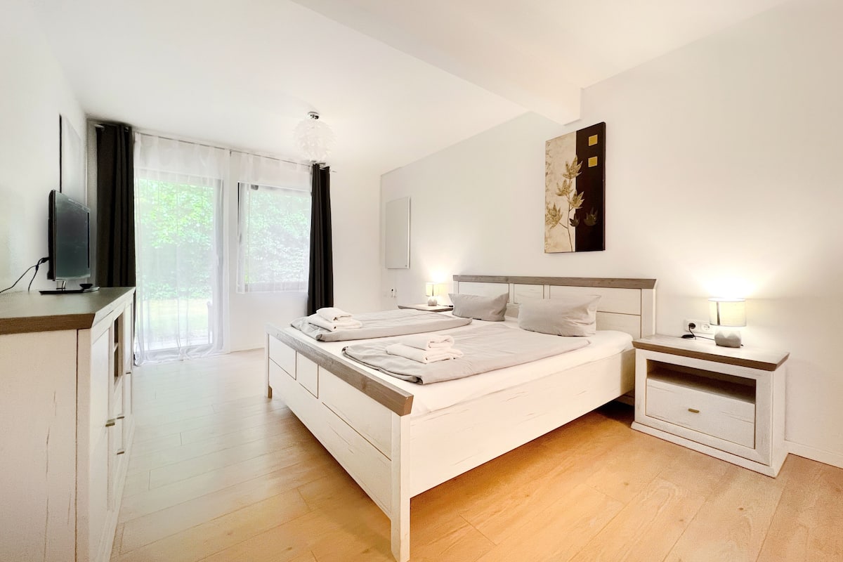 Een moderne slaapkamer met een groot bed, nachtkastjes, een flatscreen televisie en een groot raam met gordijnen.