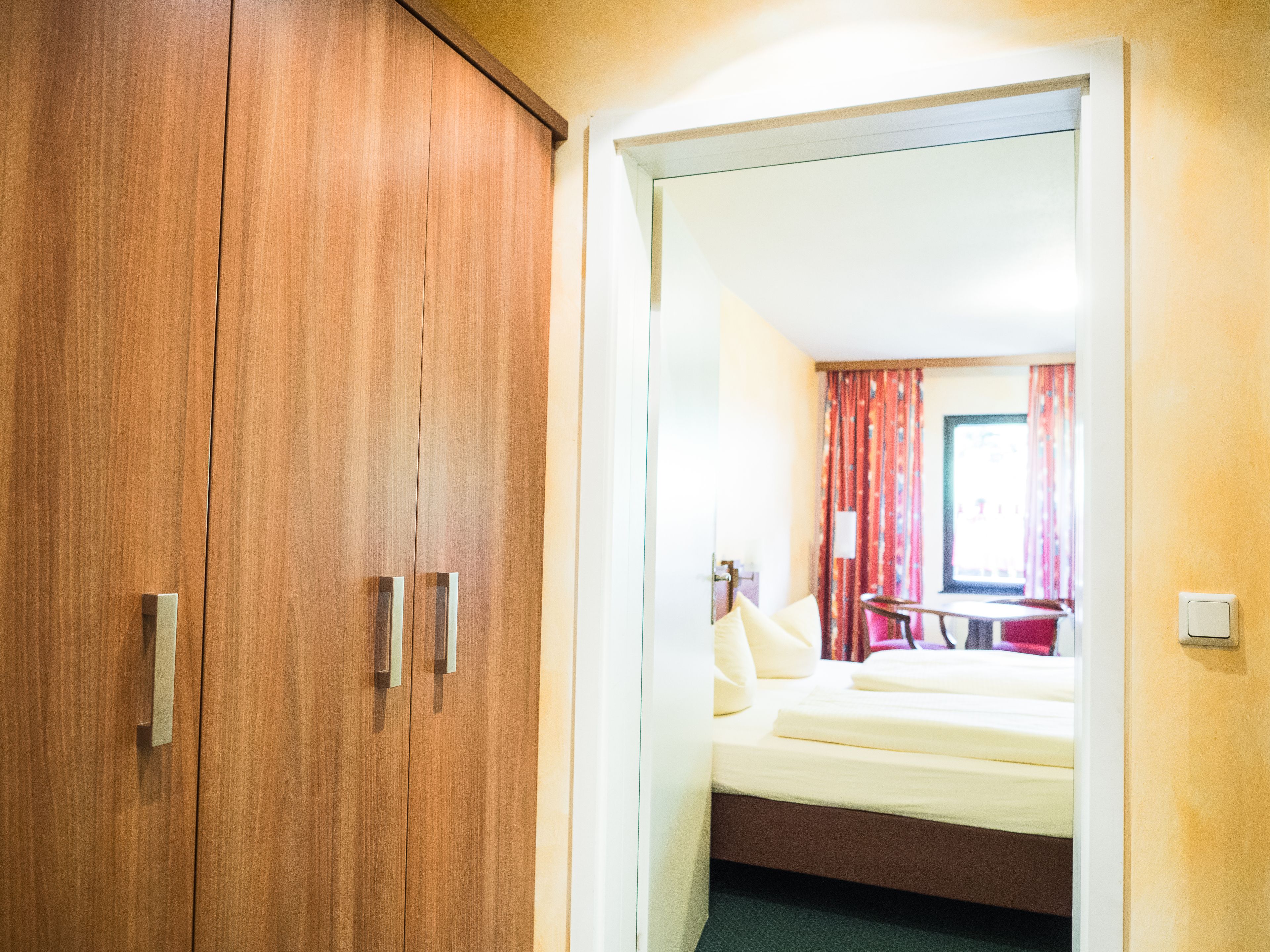 A hotel room with a wooden wardrobe, a bed with white bedding, and a window with red curtains.