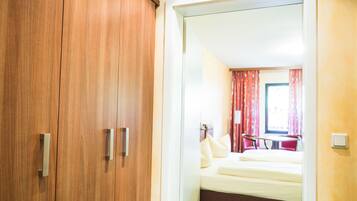 A hotel room with a wooden wardrobe, a bed with white bedding, and a window with red curtains.