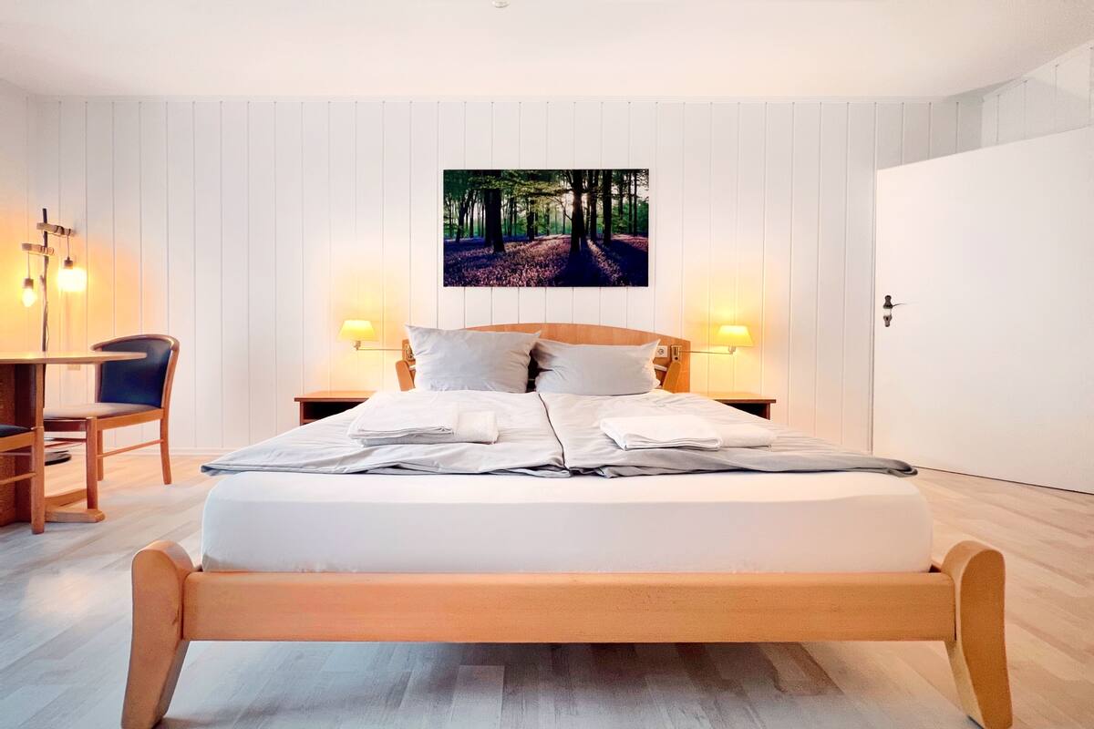 Een moderne slaapkamer met een houten bed, twee bedlampen, een bureau met stoel, en een aan de muur hangend beeld van een bospad.