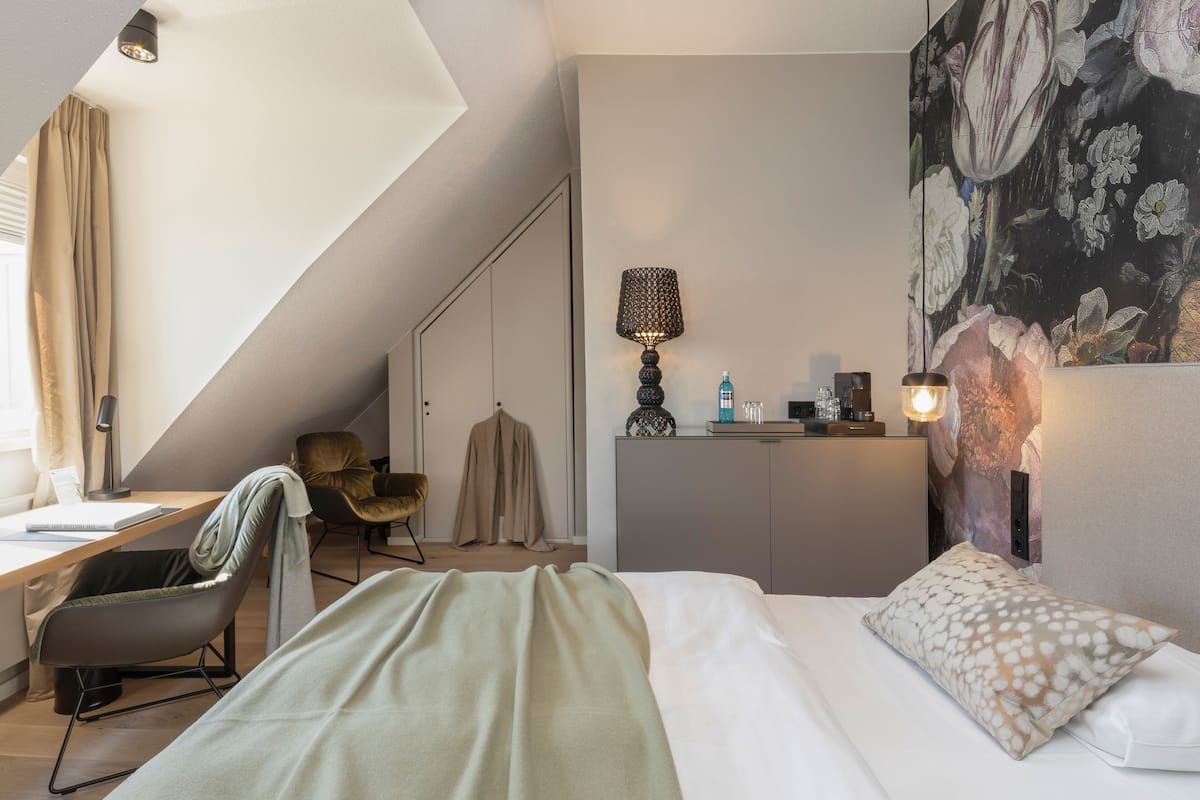 A modern hotel room with a bed, a desk, a chair, a lamp, and a wardrobe.