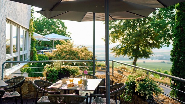 Eine Terrasse im Freien mit einem Tisch für zwei Personen, umgeben von Korbstühlen, unter einem großen Sonnenschirm.