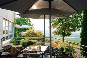 An outdoor patio with a table set for two, surrounded by wicker chairs, under a large umbrella.