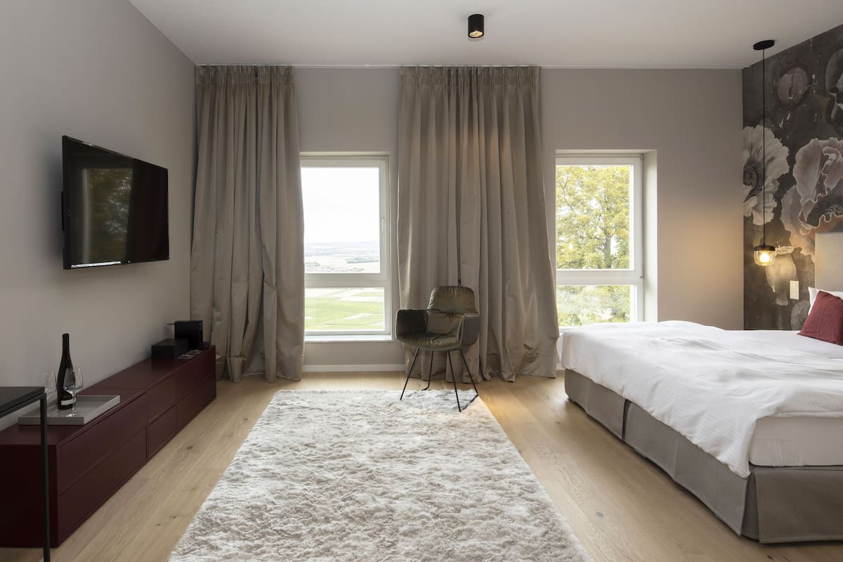 A modern bedroom with a large bed, a TV mounted on the wall, a desk with a wine bottle, and two windows with curtains.