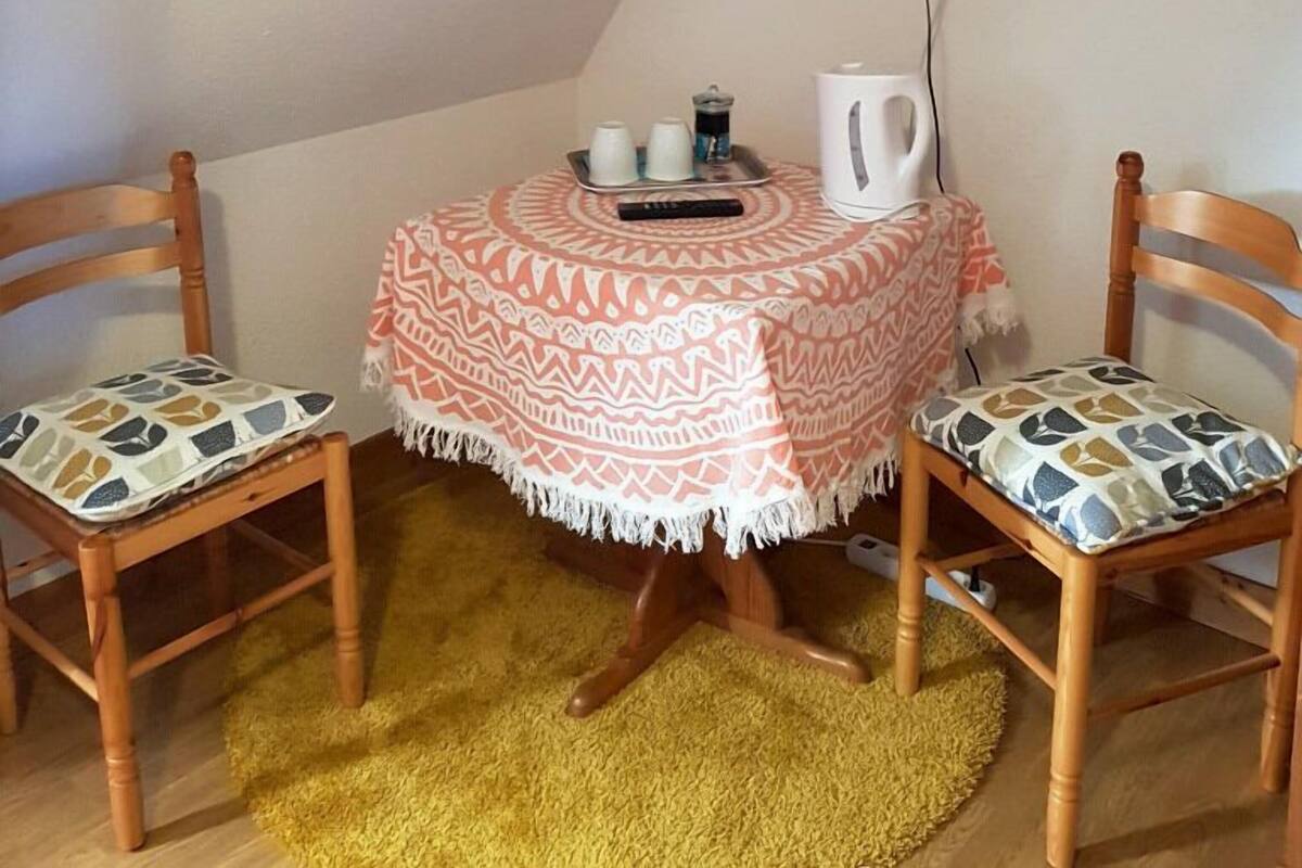 A small, cozy room with a round table covered by a patterned tablecloth, two wooden chairs with patterned cushions, a television mounted on the wall, and a yellow rug on the floor.