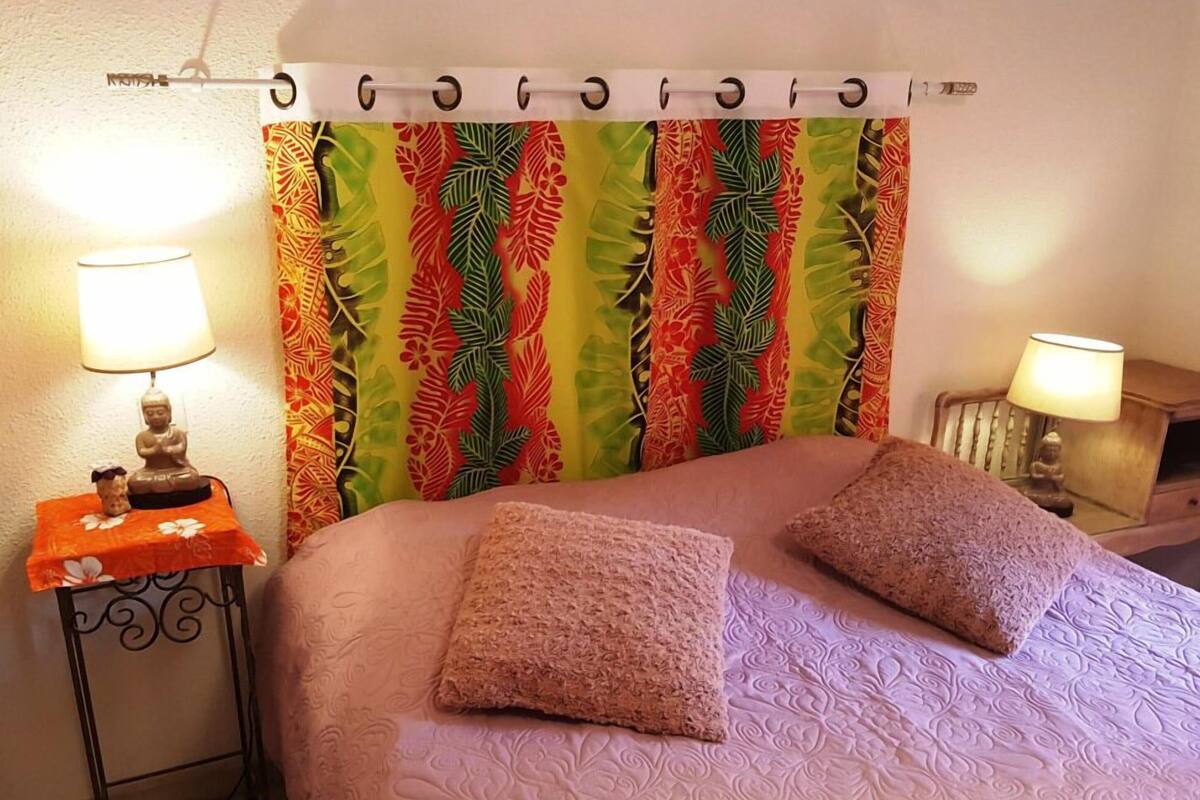 A bed with a patterned headboard, two pillows, a nightstand with a lamp, and a wall-mounted curtain.