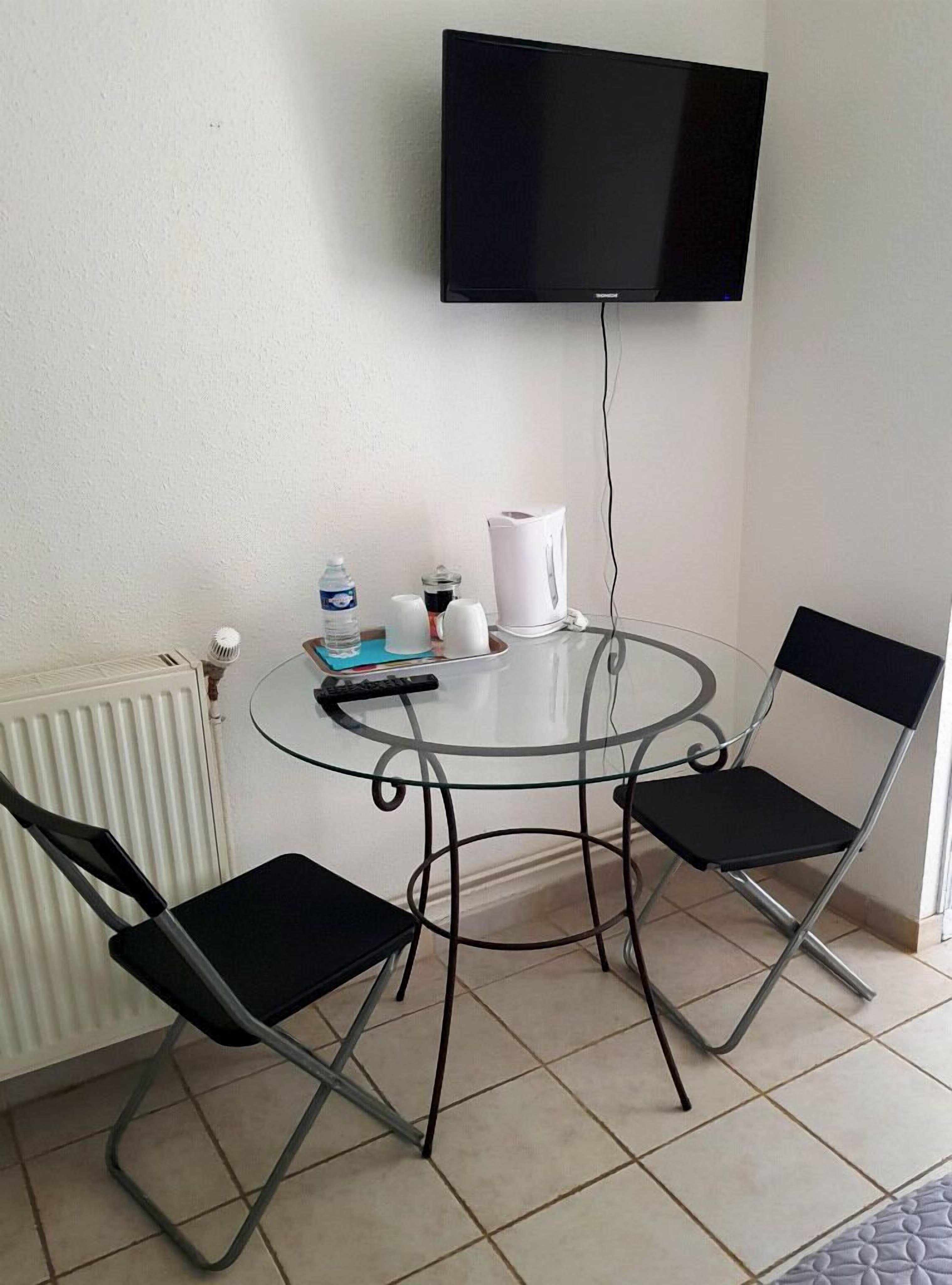 Una habitación pequeña con una mesa redonda de vidrio, dos sillas negras, un radiador blanco, una tetera blanca, una botella de agua, un libro y un televisor de pantalla plana montado en la pared.