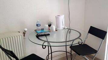 Una habitación pequeña con una mesa redonda de vidrio, dos sillas negras, un radiador blanco, una tetera blanca, una botella de agua, un libro y un televisor de pantalla plana montado en la pared.