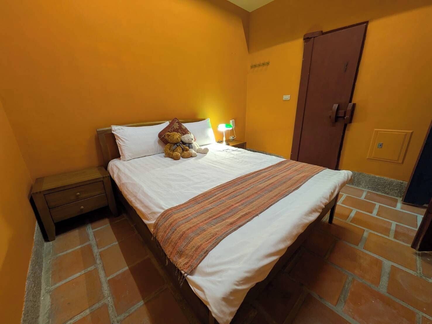A hotel room with a bed, a nightstand, a teddy bear, a lamp, and a door.