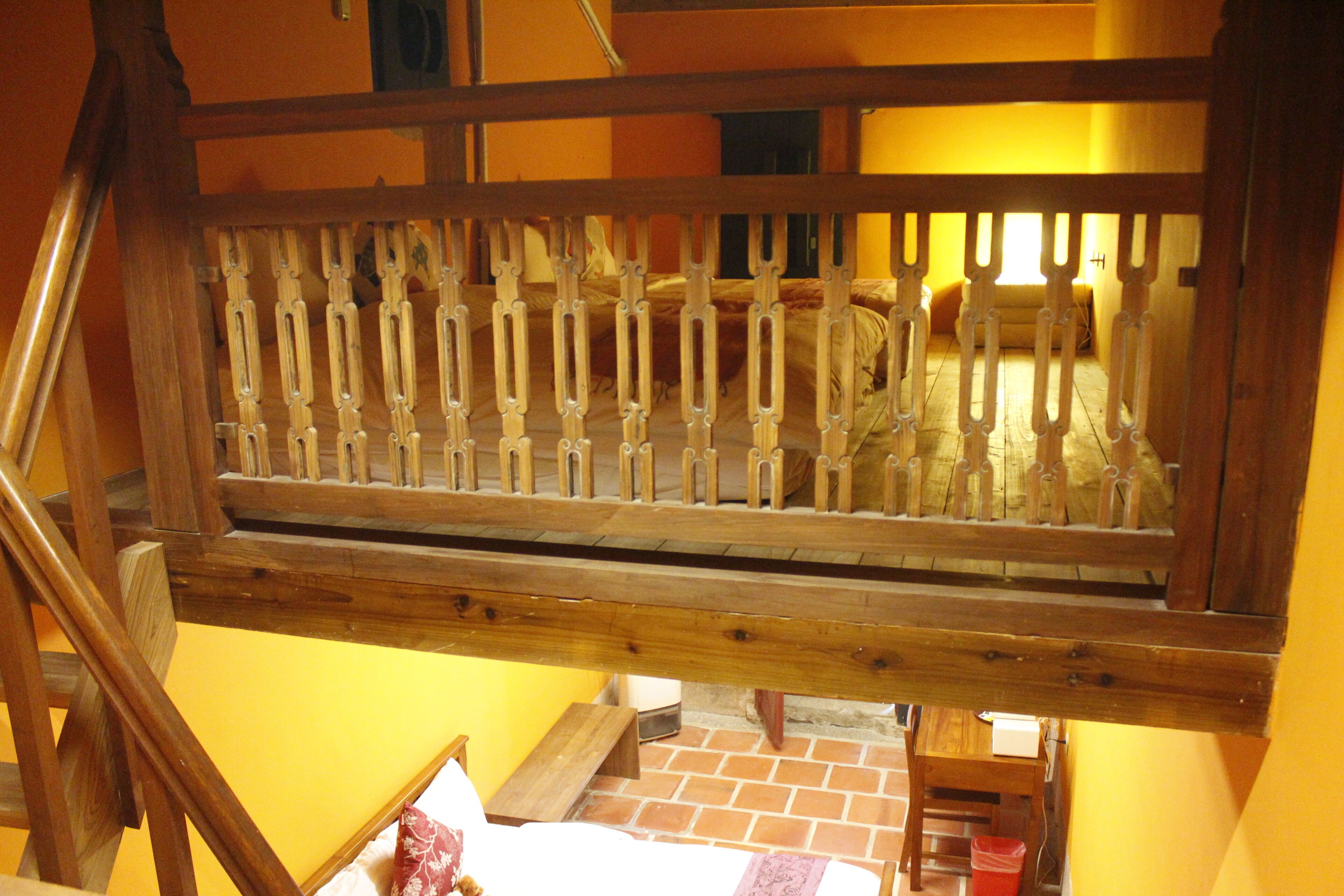 A wooden staircase with a railing, a bed with a white mattress and a red blanket, a small wooden table, and a trash bin.