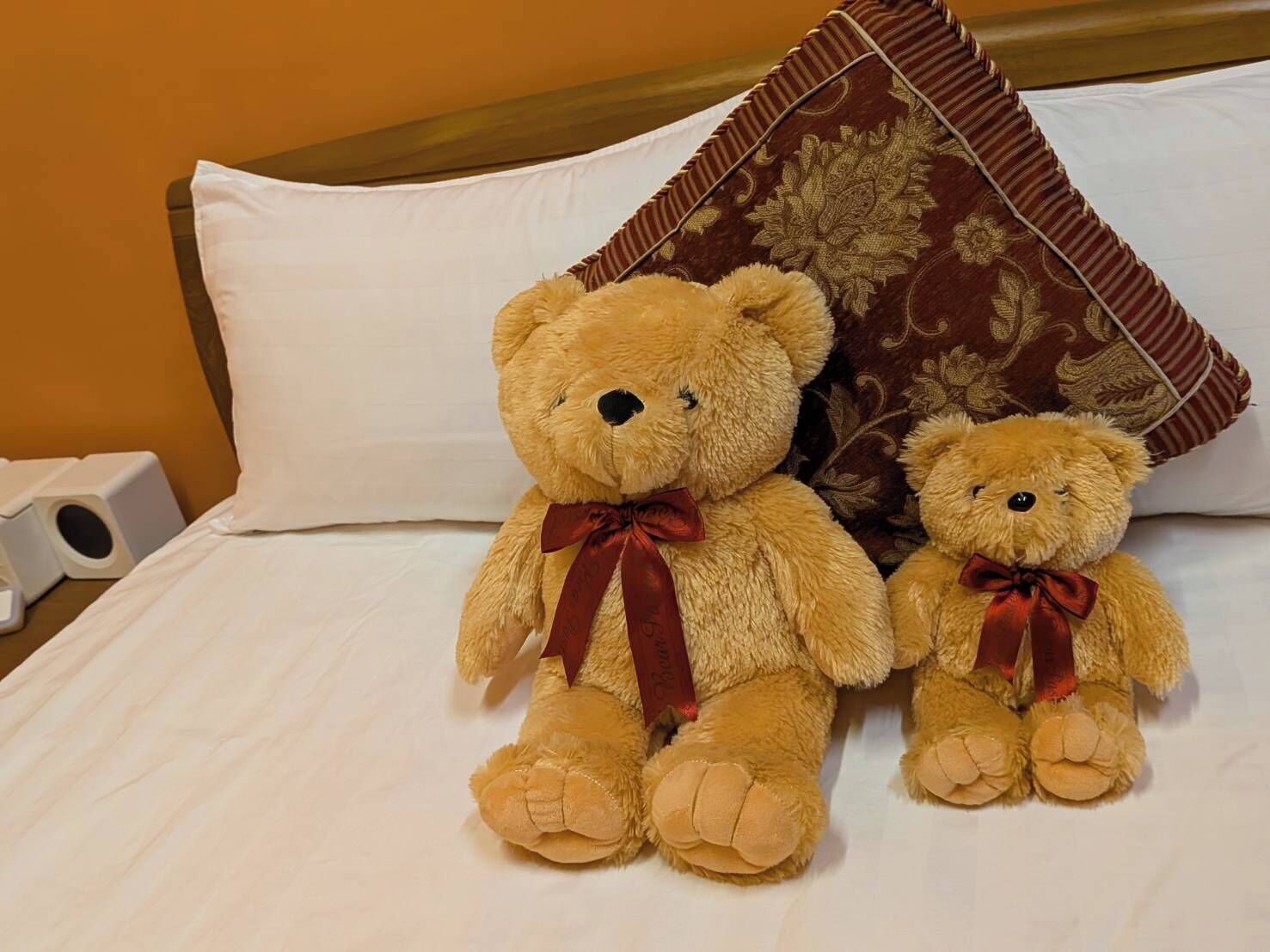 Two teddy bears on a bed with white bedding and a brown patterned pillow.
