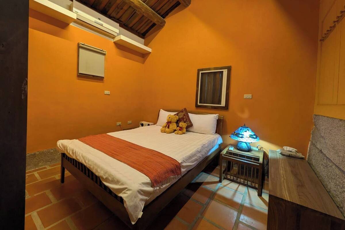 Une chambre avec un plafond en bois, un lit recouvert d’une couverture blanche et orange, un ours en peluche sur le lit, une table de chevet avec une lampe et une petite table avec un téléphone.