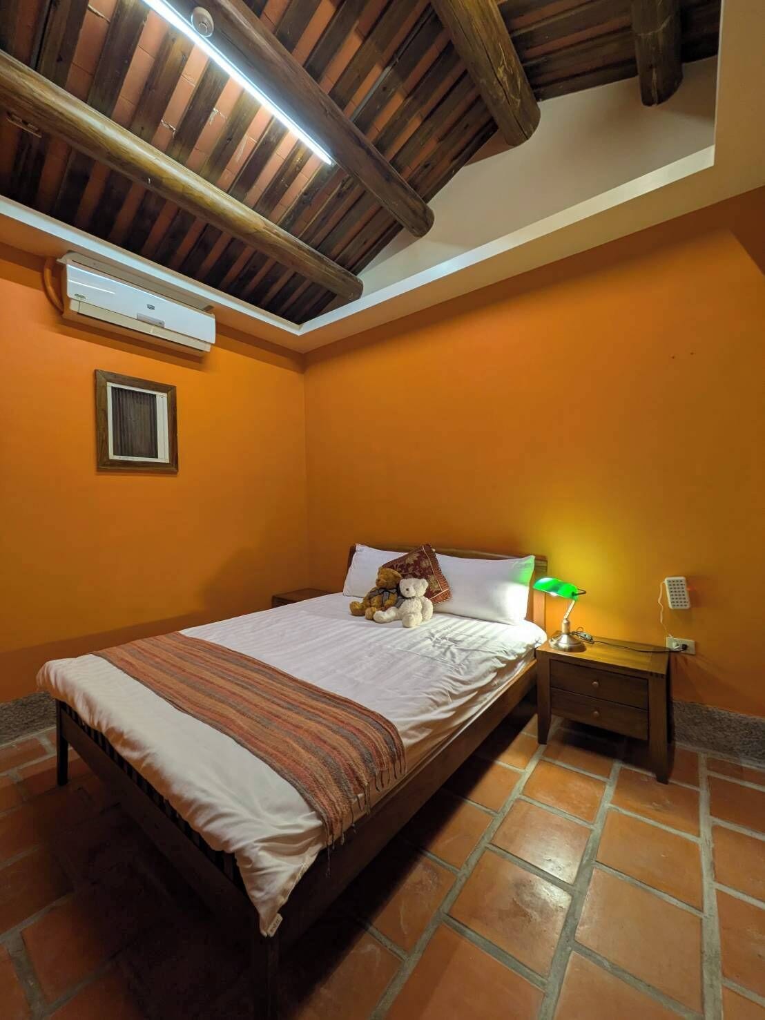 Un dormitorio con cama, mesita de noche, lámpara, un osito de peluche y una ventana.