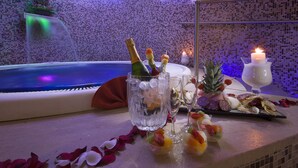 Ein luxuriöser Wellnessbereich mit einer Wand aus Mosaiksteinen, einer Whirlpool-Badewanne und einem Tisch, gedeckt mit Champagner, Snacks und einer Kerze.