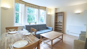 A dining area with a wooden table set for four, a grey sofa, and a bookshelf. - CityGate Farringdon (London)
