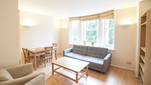 A modern living room with a sofa, dining table, and chairs. There is a window with curtains and a view of greenery outside. - CityGate Farringdon (London)