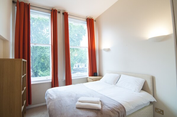 Quarto com cama, mesa de cabeceira, guarda-roupa e janela com cortinas.