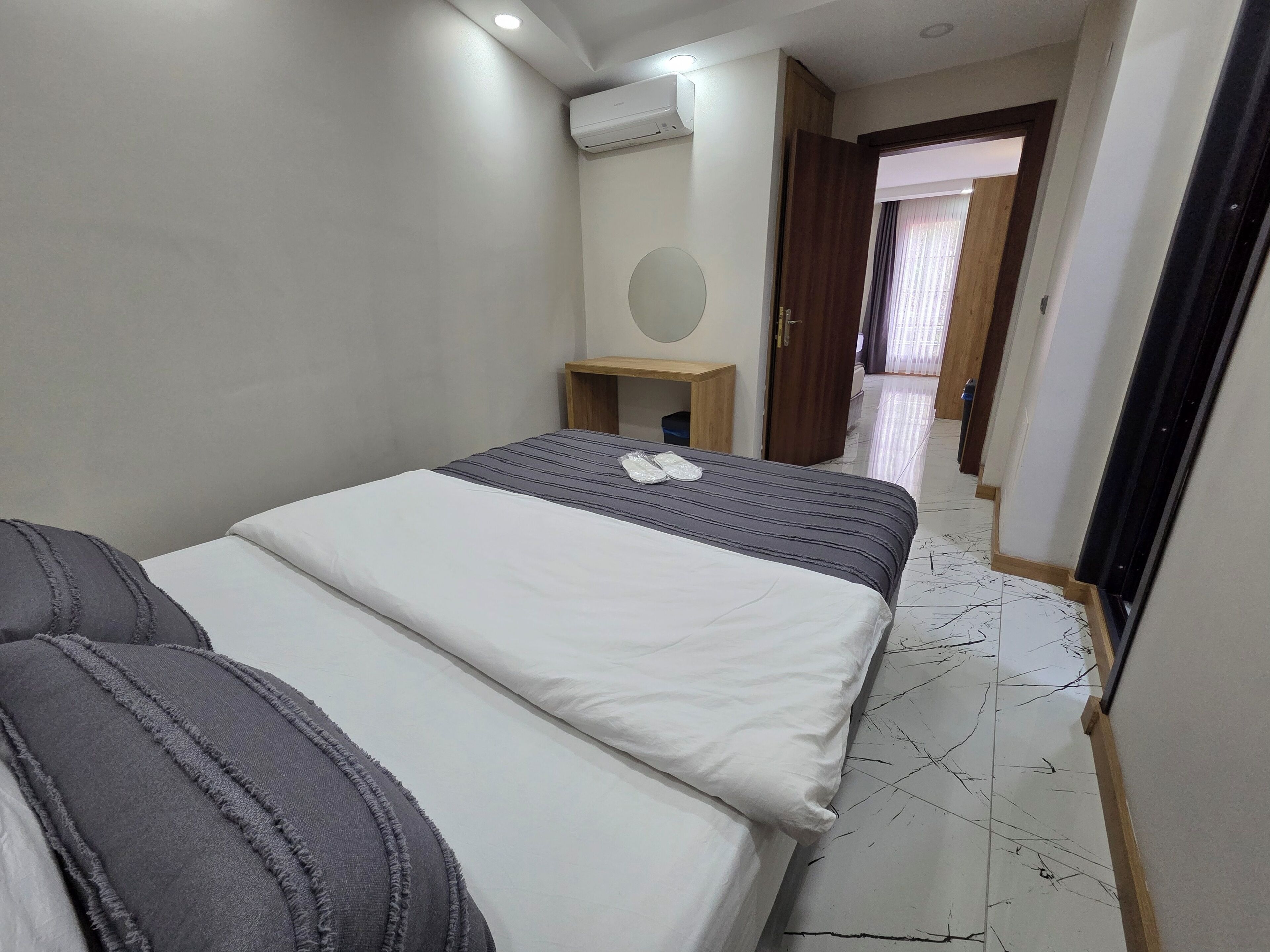 A hotel room with a bed, a bedside table, an air conditioning unit, and a view of the hallway.