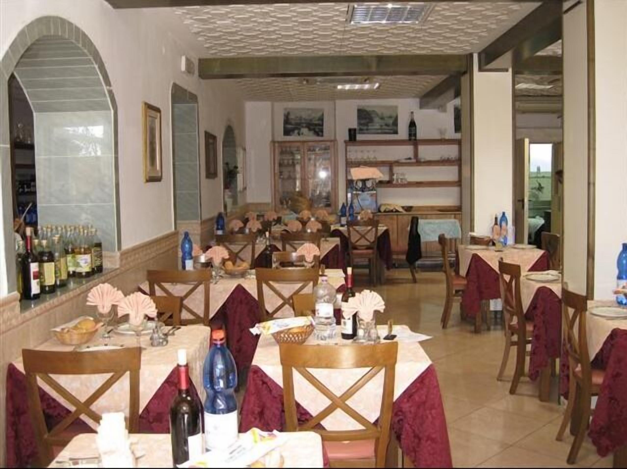 A restaurant with multiple tables and chairs, some with red tablecloths. There are bottles and glasses on the tables, and a large window in the background.