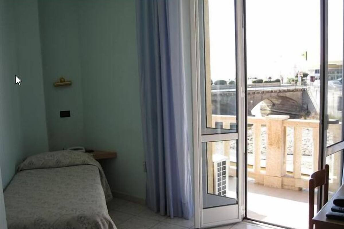 A room with a bed, a window with curtains, and a view of a balcony with a railing.