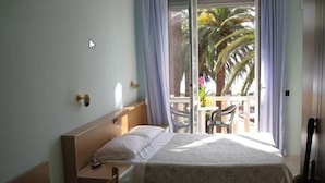 Habitación de hotel con cama, escritorio y silla. Tiene un balcón con vistas a palmeras.