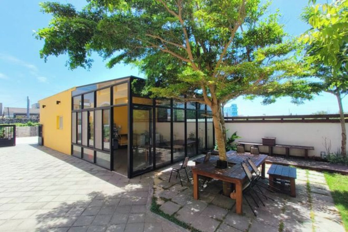 A modern building with glass walls, a patio area with a wooden table and chairs, and a tree providing shade.