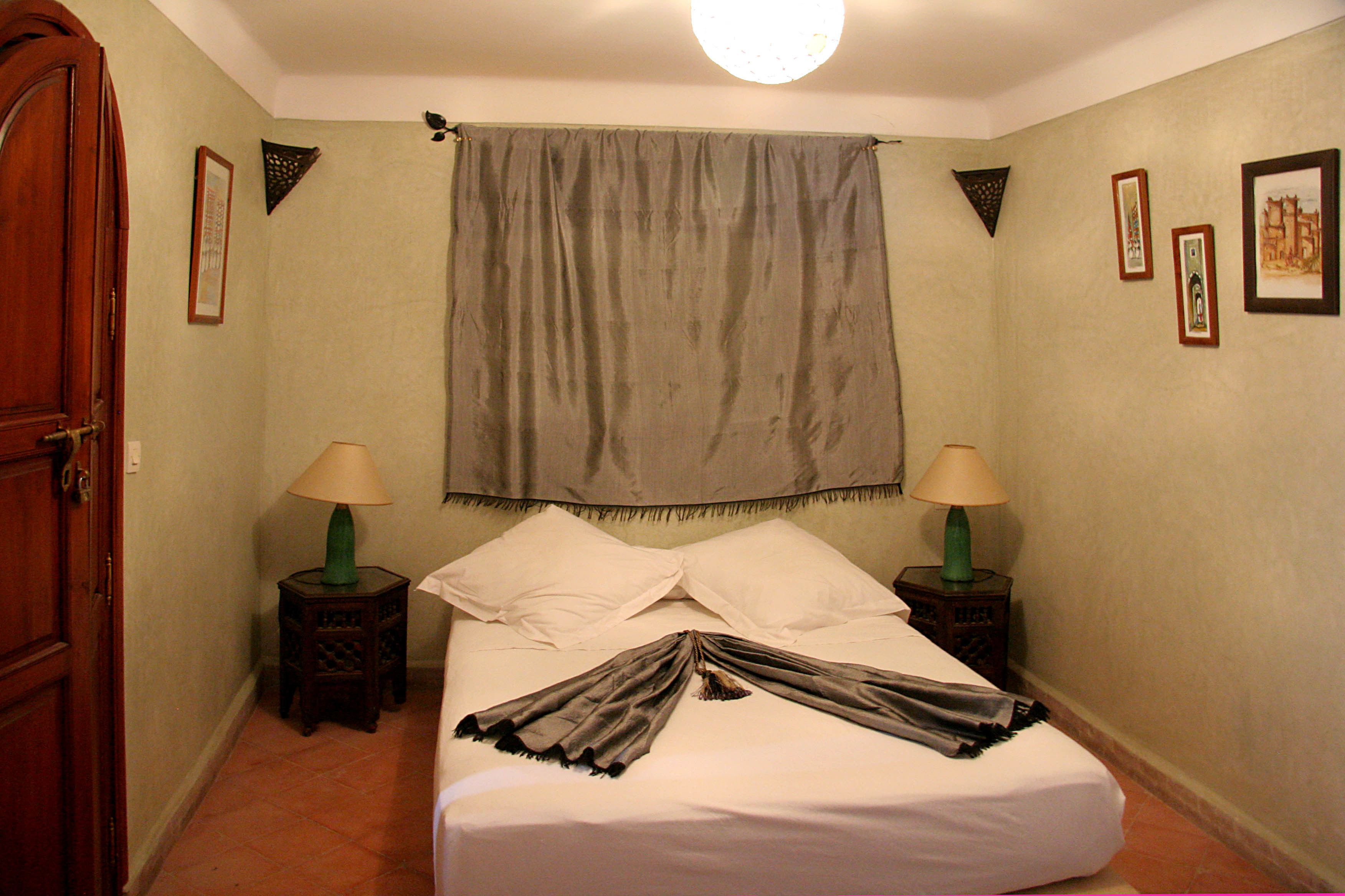 A hotel room with a bed, two nightstands, a lamp on each, a large window with a curtain, and a door.