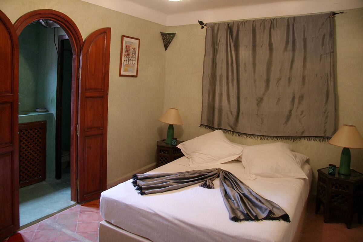 A bedroom with a bed, two nightstands, a wooden wardrobe, a door, a curtain, and a framed picture on the wall.