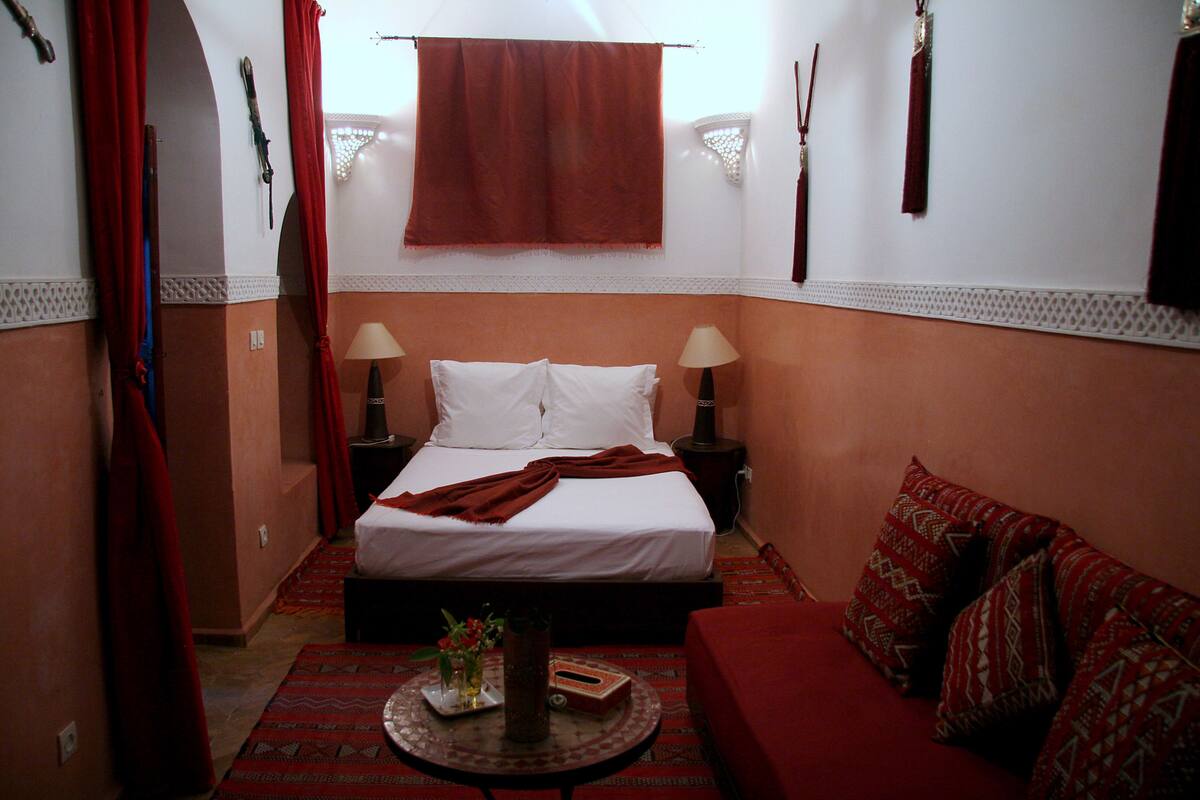 A hotel room with a bed, two bedside tables, a red sofa, and a small round table with a vase and book.