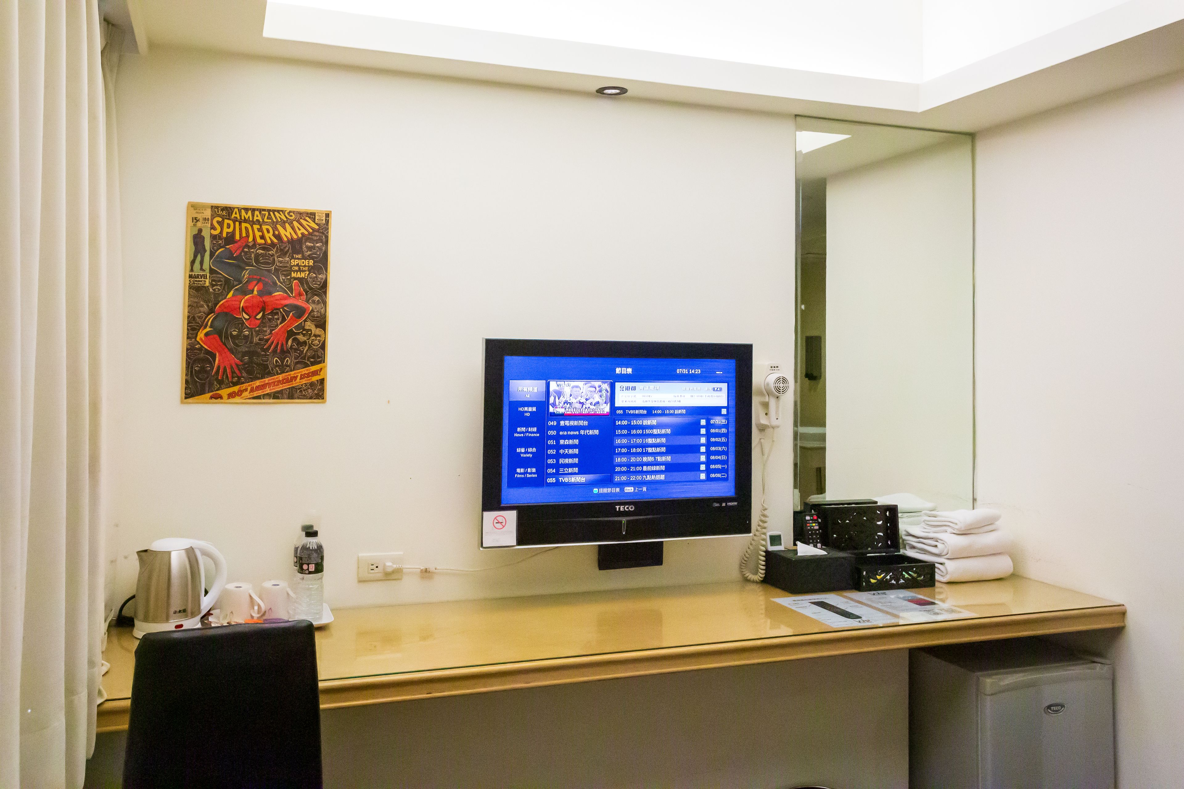 A hotel room with a desk, a flat-screen TV, a poster, a coffee maker, a bottle of water, and a trash bin.