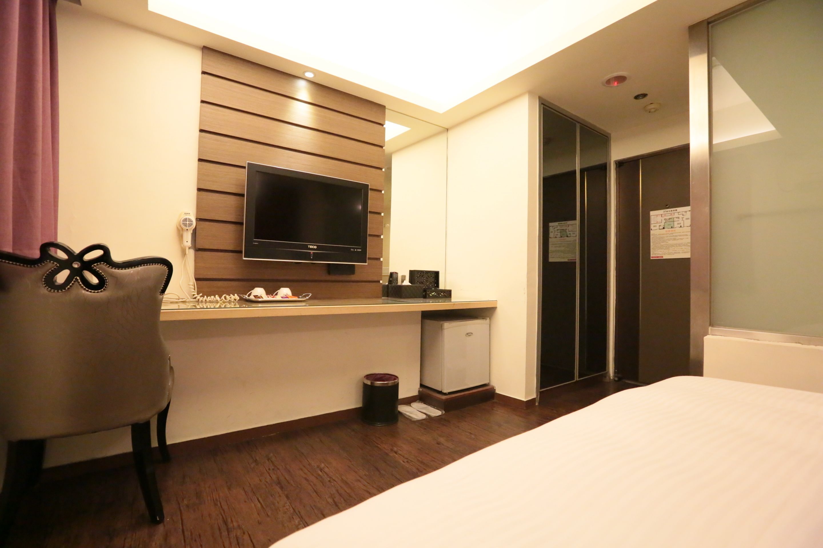 A hotel room with a bed, a desk with a flat-screen TV, a chair, a small refrigerator, and a closet.