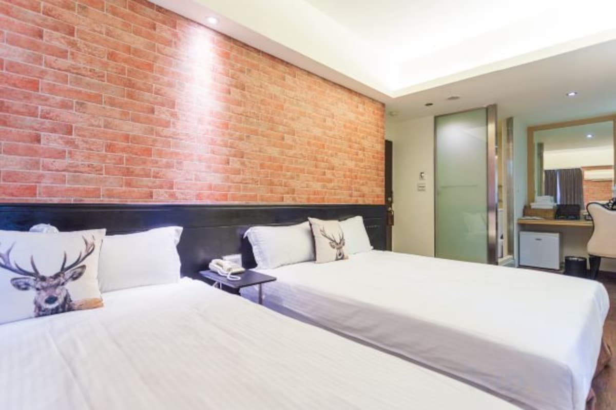 A hotel room with two beds, a brick accent wall, a desk with a chair, and a mirror.