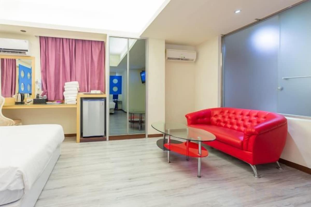 A hotel room with a bed, a red sofa, a small table, a desk with a computer, and a mirror.